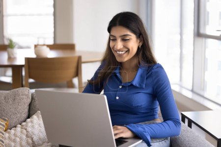 Photo for Happy young Indian freelance employee woman working at home, typing on laptop, sitting on couch, holding pc on lap, chatting online, smiling, laughing, enjoying remote communication on Internet - Royalty Free Image