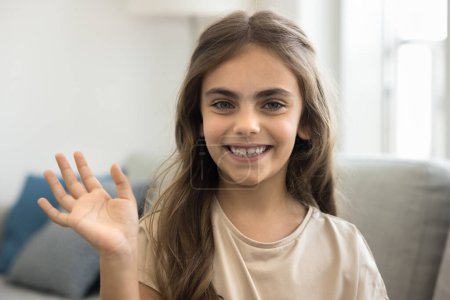 Photo for Happy cute little blogger kid girl with long hair and bushy brows waving greeting hand at camera, saying hello, smiling, laughing, speaking on online streaming, video call, enjoying communication - Royalty Free Image