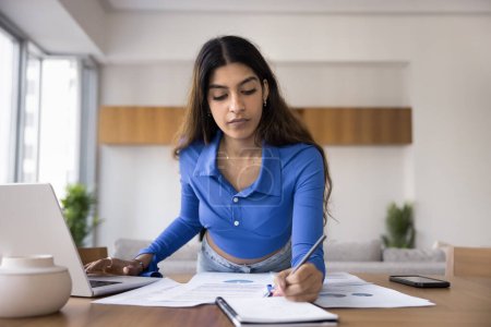 Photo for Busy young Indian business professional woman working on project at home, studying paper documents, marketing reports, typing on laptop computer at table, writing notes - Royalty Free Image