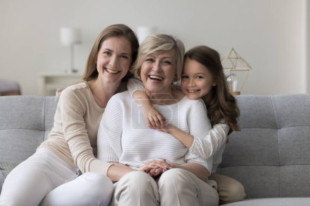Photo for Three female generations family portrait. Cute little girl hugging tightly her beloved granny sit on sofa with young mom, smile look at camera, enjoy leisure time together. Unconditional love and ties - Royalty Free Image