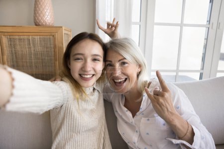 Middle-aged mum and pre-teen daughter hold device, smile, show hand gestures looking at camera, record video for social media, take selfie pictures, webcam view. Videocall, fun use modern technology