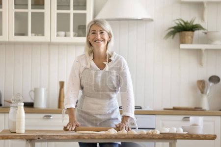Photo for Portrait of happy mature woman wear apron cook alone in modern domestic kitchen, flattening dough with rolling-pin for sweet homemade pastry, enjoy cookery process on weekend at home. Chores, cooking - Royalty Free Image