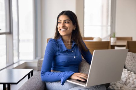 Photo for Cheerful positive young Indian woman working on freelance project at home, sitting on couch, typing on laptop, looking away, thinking on creative ideas, Internet startup, online education - Royalty Free Image