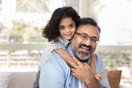 Photo for Happy loving Indian grandfather piggybacking sweet beloved preschool granddaughter at home, sitting on couch, enjoying kids hug, looking at camera, smiling, laughing - Royalty Free Image