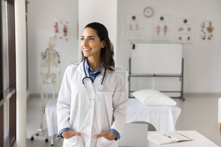 Cheerful beautiful young Latin doctor woman with stethoscope on neck looking at window away with toothy smile, thinking. Happy medical professional, intern posing in hospital anatomy boardroom