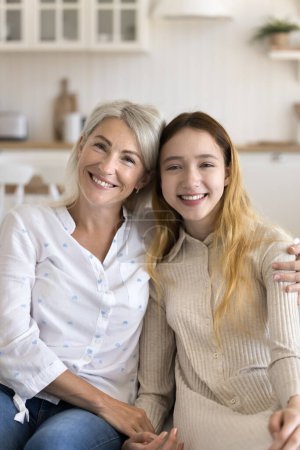 Vertical portrait of beautiful middle-aged mother cuddling her pretty pre-teen daughter sitting on couch, having pleasant leisure together, enjoy their good, trustful relationships spend time at home