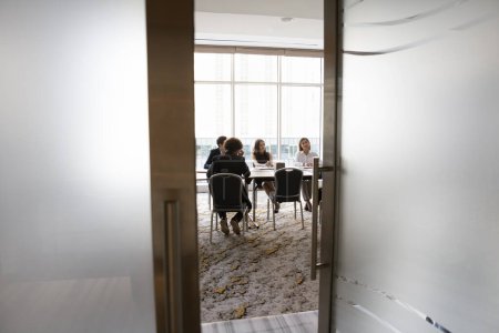 Photo for Diverse young business team sitting at large table in meeting room, talking, discussing collaboration on project, brainstorming on creative ideas. Full length candid shot, door view - Royalty Free Image