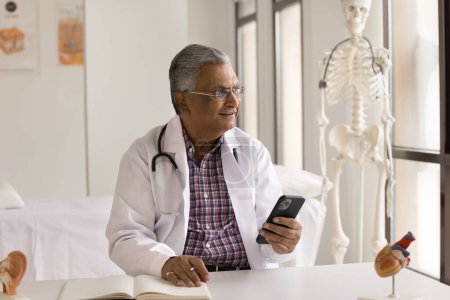 Photo for Positive thoughtful older medical practitioner in white coat using mobile phone for remote communication, at workplace table, looking away, thinking on online consultation to patient - Royalty Free Image