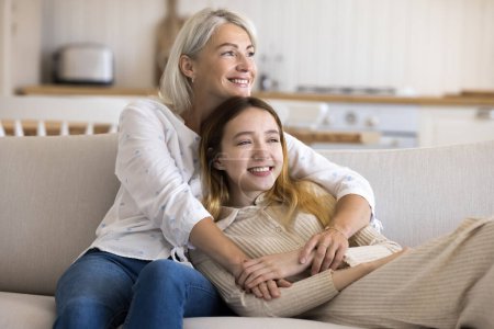 Photo for Cheerful adolescent girl spends leisure time with mature mother at home, sit resting on sofa together, enjoy tender moments, show love and care. Happy family of two generations hugging and daydreaming - Royalty Free Image
