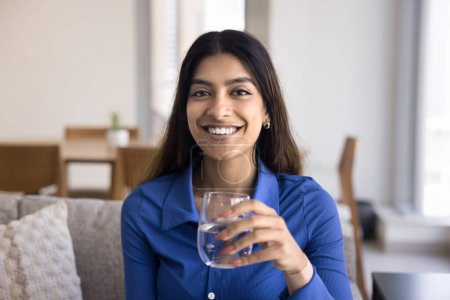 Foto de Mujer india positiva hermosa y saludable bebiendo agua clara, sosteniendo el vaso con bebida fría, cuidando la salud, la nutrición, el equilibrio acuático, la hidratación, mirando a la cámara con una sonrisa dentada - Imagen libre de derechos