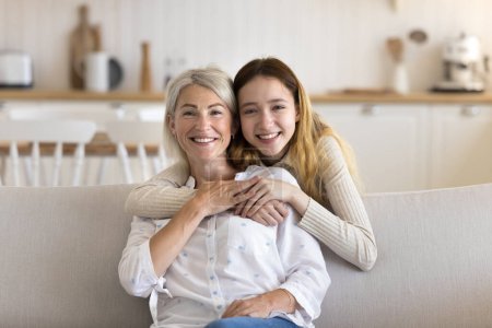 Photo for Two generations females enjoy carefree family leisure time at home, smiling, looking at camera. Happy pre-teen daughter embraces mature mother from behind with love, demonstrate care and tenderness - Royalty Free Image
