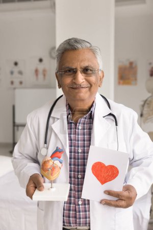 Photo for Happy elderly Indian doctor man holding human heart model and greeting card at chest, looking at camera, smiling. Successful mature cardiologist in white coat and glasses vertical portrait - Royalty Free Image