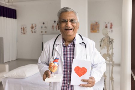 Foto de Alegre médico cardiólogo indio viejo posando en la oficina del hospital, sosteniendo el modelo de órgano del corazón humano, mirando a la cámara, sonriendo, riendo. Profesional médico promoviendo chequeo de salud - Imagen libre de derechos