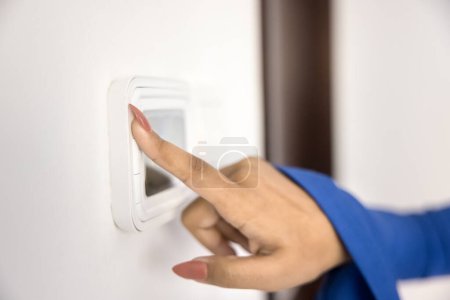 Young woman using climate control panel at home, regulating thermostat, air temperature in apartment, pressing button on wall mounted device with display. Close up of hand and smart home gadget