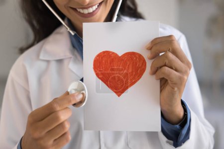 Photo for Hands of young doctor woman holding stethoscope and greeting card with drawn red heart, smiling, playing listening to heartbeat, offering cardiac medical checkup, examination. Close up cropped shot - Royalty Free Image