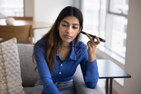 Photo for Serious focused young adult Indian girl listening to voice audio message on telephone, holding smartphone dynamic at ear, thinking, looking away in deep thoughts - Royalty Free Image