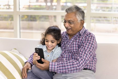 Téléchargez les photos : Heureux grand-père indien et mignon petit enfant petite-fille regarder contenu drôle sur téléphone mobile, en utilisant l'application en ligne ensemble, assis, étreignant sur le canapé à la maison, tenant appareil numérique - en image libre de droit