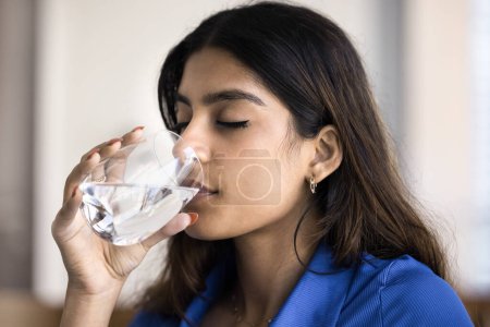 Téléchargez les photos : Belle jeune fille indienne des années 20 avec les yeux fermés profiter de l'eau pure fraîche, boire une boisson froide à partir du verre, satisfaire la soif, garder l'équilibre d'hydratation, peau faciale lisse et saine - en image libre de droit