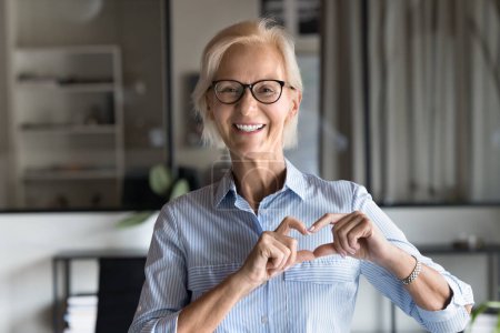 Photo for Cheerful senior blonde business lady making hand heart gesture, joining fingers at chest, expressing love, support, kindness, looking at camera with toothy smile for office portrait - Royalty Free Image