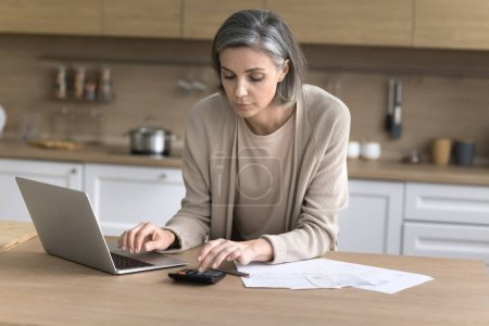 Photo for Focused middle aged woman check family budget standing at kitchen table, make e-bank on-line payment on laptop, calculates bills, do bookkeeping job, counting incomes and expenses. Accounting, savings - Royalty Free Image