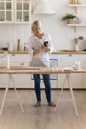 Téléchargez les photos : Sourire femme au foyer d'âge moyen debout à la table de cuisine avec smartphone, vérifier la recette en ligne, prendre des photos du processus de cuisson sur téléphone mobile, bavarder, profiter du temps de cuisson à la maison. Technique moderne, culinaire - en image libre de droit