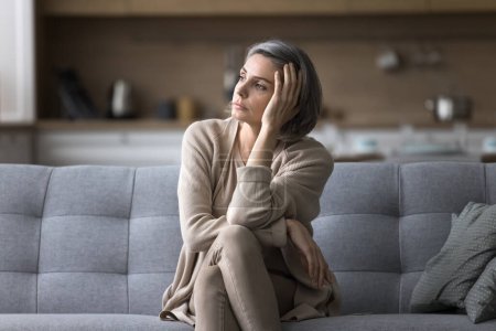 Attractive silent middle-aged woman relaxing seated on couch in living room, staring out window, ponders, missing, deep in thoughts, looks pensive and thoughtful, spend boring weekend alone at home