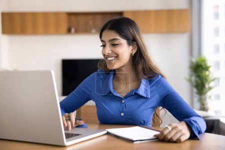Photo for Cheerful young Indian student girl watching educational webinar, online training lecture on laptop, writing notes, speaking on learning Internet conference meeting, using computer - Royalty Free Image