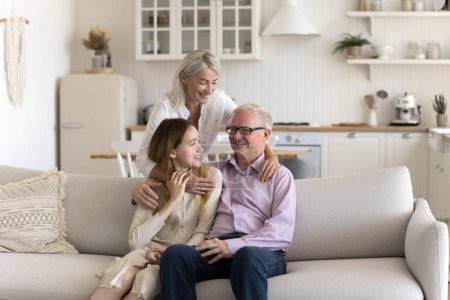 Friendly family have conversation sitting on sofa, senior man relax with mature daughter and adolescent granddaughter in living room, enjoy spending time together. Harmony, relationship, understanding