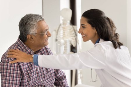 Photo for Cheerful young doctor woman touching shoulder of senior Indian patient with support, care, talking to man, laughing, telling good medical news about health, healthcare, therapy result - Royalty Free Image