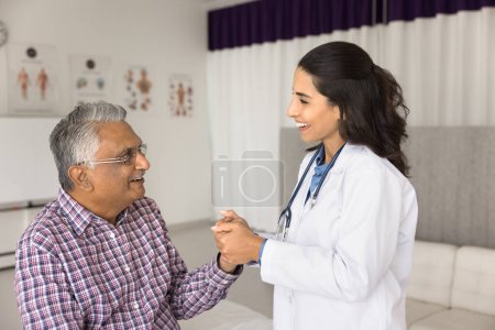 Feliz joven médico empático tocando la mano del anciano paciente indio con enfermedades geriátricas, sosteniendo el brazo con apoyo, calmando al hombre mayor, dando atención médica, consulta, sonriendo, riendo