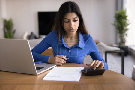 Photo for Serious young Indian accountant woman calculating domestic expenses, typing on calculator at laptop, paying bills, checking paper invoices. Freelancer working at home, counting budget - Royalty Free Image