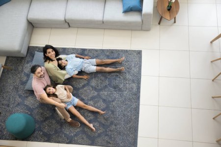 Photo for Happy middle aged Latin couple of parents and two kids resting on carpeted warm floor, looking up at camera, smiling, laughing, enjoying home family leisure. Top view, full length shot - Royalty Free Image