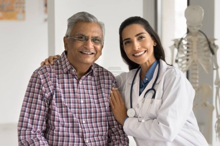 Positiv schöne junge Medizinerin umarmt ältere indische Patientin, drückt Wärme, Fürsorge, Unterstützung, Empathie aus, blickt mit zahmem Lächeln in die Kamera, posiert für ein Porträt