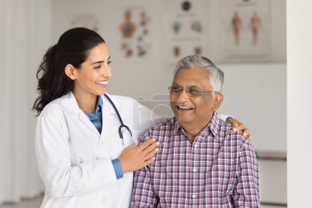 Foto de Mujer geriatra alegre y vieja paciente india riendo en la clínica juntos, hablando, discutiendo buenas noticias, terapia exitosa, tratamiento. Doctor tocando hombros mans con cuidado - Imagen libre de derechos