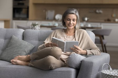 Photo for Untroubled smiling mature woman spend leisure time at home alone, settled down on comfortable sofa with interesting paper book, read favorite literature, enjoy peaceful weekend with bought bestseller - Royalty Free Image