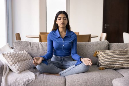 Photo for Focused tranquil young Indian yogi girl meditating at home, sitting on couch, keeping zen mudra, enjoying practice, calmness, peace, training focus, concentration, doing breath work - Royalty Free Image
