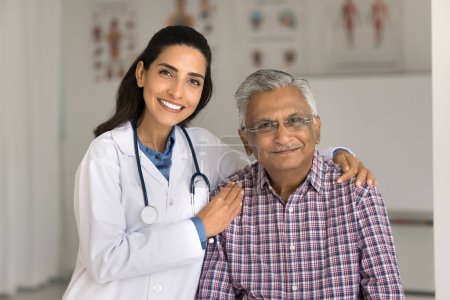 Photo for Happy beautiful young Latin doctor woman embracing senior Indian patient man, touching mans shoulders with care, support, looking at camera, smiling, posing for portrait in hospital office - Royalty Free Image