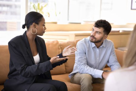 Photo for Serious multiethnic male and female colleagues discussing work tasks at corporate networking meeting. Confident young African businesswoman talking to coworker man on office couch - Royalty Free Image