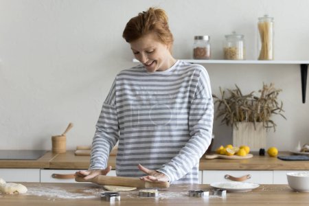 Photo for Smiling young woman cooking alone in modern kitchen, use rolling-pin flattening dough for homemade sweet dessert, prepare cookies enjoy cook process on weekend at home. Hobby, chores, food preparation - Royalty Free Image