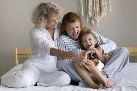 Little 6s grandkid sit on bed enjoy playtime with young 30s mother and older 55s grandma. Mother and granny tickling adorable preschooler kid girl spend time together at home. Family ties, connection