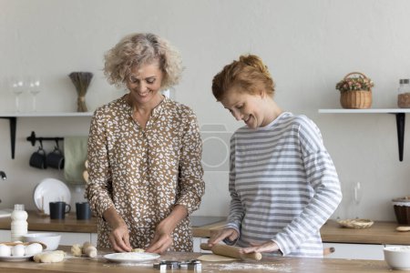 Photo for Smiling older and younger women cooking together in the kitchen. Happy mature female enjoy food preparation with young adult daughter, standing at table flattening homemade dough make cookies at home - Royalty Free Image
