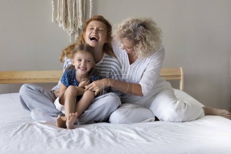 Téléchargez les photos : Deux générations de femmes caucasiennes, mamie plus âgée et jeune maman partagent un moment chaleureux, chatouillant petite fille assise sur le lit, profiter de leurs relations amicales, amour illimité et joie sur le week-end confortable - en image libre de droit