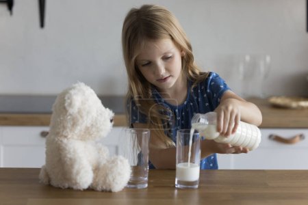 Téléchargez les photos : Petite fille tenir bouteille en plastique verse du lait dans deux verres pour elle et pelucheux ours jouet. Bonne habitude de vie pour le développement et la croissance des enfants, vitamines et minéraux nécessaires à la santé globale des enfants - en image libre de droit