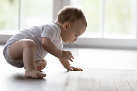 Photo for Cute little baby in bodysuit and diaper makes first steps on warm floor, close up. Adorable infant toddler crawling, try to walk in cozy living room. New generation, child growth and life exploration - Royalty Free Image