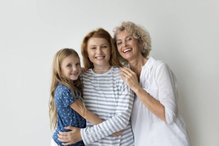 Beautiful multi-generational relative women hugging posing on gray studio background, smile look at camera enjoy their harmonic relationship, friendship and family ties, showing togetherness and unity