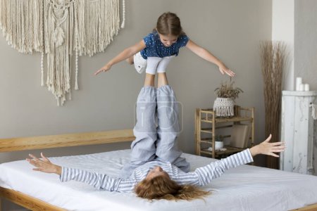 Kleines Mädchen gibt vor, in der Luft zu fliegen, genießt die Zeit mit Mutter im Bett liegend, hebt ihre süße Vorschultochter auf ausgestreckten Beinen, macht Sport, verbringt am Wochenende aktive Zeit im gemütlichen Schlafzimmer zu Hause
