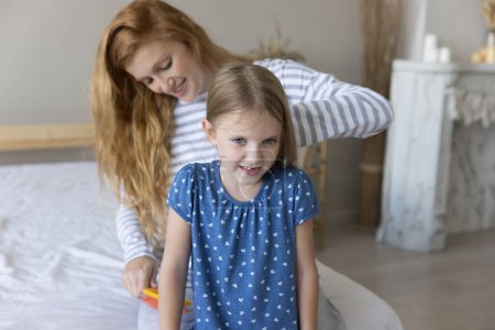 Young loving mother brushing long hair of cute little 5s daughter seated on bed in cozy bedroom, help to kid with morning routine, showing care, feeling love, enjoy talk and time together. Motherhood