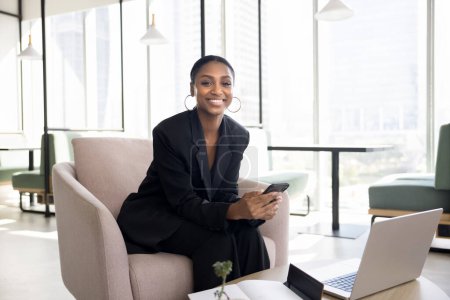 Foto de Mujer empresaria africana joven y segura de sí misma mirando a la cámara con sonrisa feliz, sentada en un sillón, trabajando en un cómodo espacio de coworking, sosteniendo el teléfono móvil. Retrato de negocios - Imagen libre de derechos