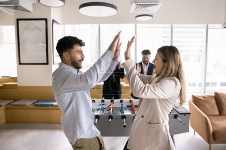 Photo for Happy couple of office workers beating colleagues in toy football, giving high five at table soccer stand, smiling, laughing, enjoying win, team success, recreation on work break - Royalty Free Image