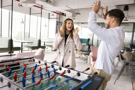 Photo for Couple of happy excited office employees giving high five over corporate board game, playing table soccer, toy football, having fun in modern co-working space with environment for recreation, activity - Royalty Free Image